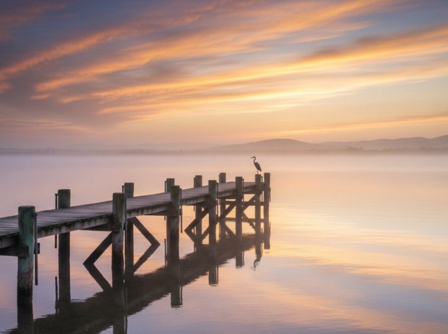 An aerial shot capturing the stunning Bittern Victoria's coastal serenity fine art photography, showcasing the dramatic confluence of a tranquil inlet with the rugged coastline at dawn, a lone fisherman casting a line, bathed in golden light, representing an epic moment of peace and natural grandeur.