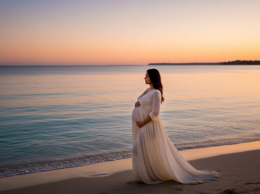 A radiant pregnant woman in a flowing white gown, silhouetted against a dramatic sunset over the turquoise waters of Safety Beach, showcasing the serene beauty of Coastal Maternity Photography Mount Martha Victoria.