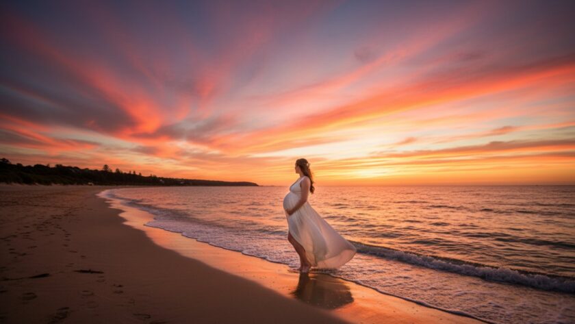 An epic moment from a dreamy Mornington sunset maternity photoshoot, featuring a glowing pregnant woman silhouetted against a vibrant orange and purple sky on the beach, with gentle waves at her feet.