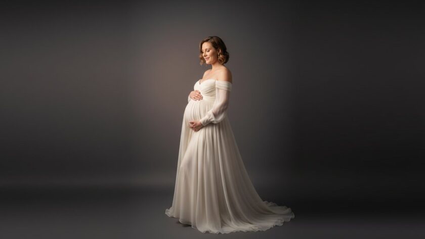 A serene and elegant portrait featuring a radiant pregnant woman in a flowing gown, bathed in soft studio light, embodying the timeless Safety Beach studio maternity photography experience.