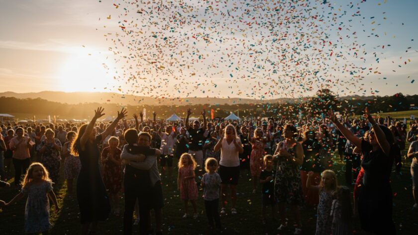 A wide shot capturing the vibrant Tremont Victoria outdoor event photography, featuring a dynamic group of people cheering enthusiastically amidst the lush greenery of a local park at sunset, conveying pure joy and connection.