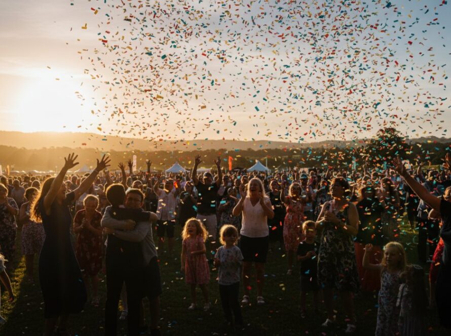 A wide shot capturing the vibrant Tremont Victoria outdoor event photography, featuring a dynamic group of people cheering enthusiastically amidst the lush greenery of a local park at sunset, conveying pure joy and connection.