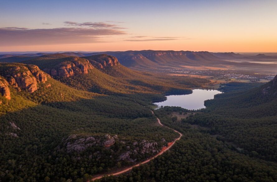 An epic drone shot capturing the vast, stunning landscape of the Grampians National Park near Ararat, Victoria, Australia, showcasing Ararat drone photography services for breathtaking regional views. The sun rises over jagged peaks, casting long shadows, with a winding road leading towards the distant town, evoking a sense of adventure and grandeur.