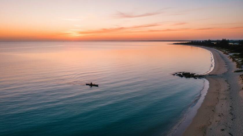 An breathtaking aerial photograph capturing the Aspendale drone photography coastal beauty at sunrise, showing the golden light illuminating the sandy beach and calm turquoise waters, with a lone kayaker gliding through the reflections, taken from a high vantage point.