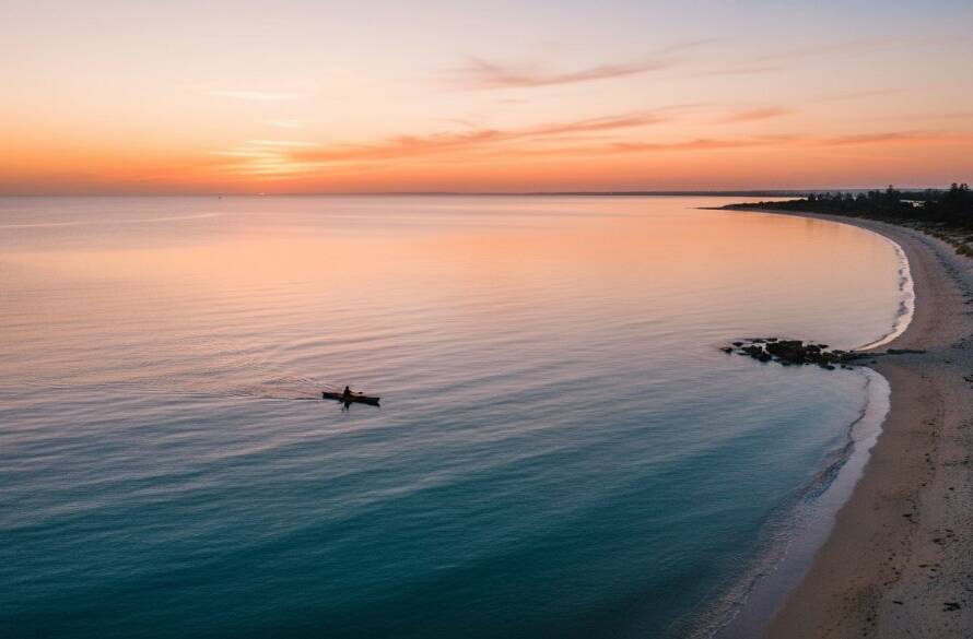 An breathtaking aerial photograph capturing the Aspendale drone photography coastal beauty at sunrise, showing the golden light illuminating the sandy beach and calm turquoise waters, with a lone kayaker gliding through the reflections, taken from a high vantage point.