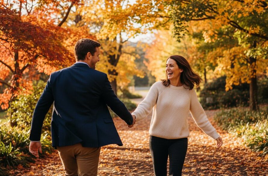 Two people laughing joyfully while walking hand-in-hand through the autumn leaves near Lake Daylesford, illuminated by golden hour light, perfectly capturing authentic Daylesford candid photography moments.