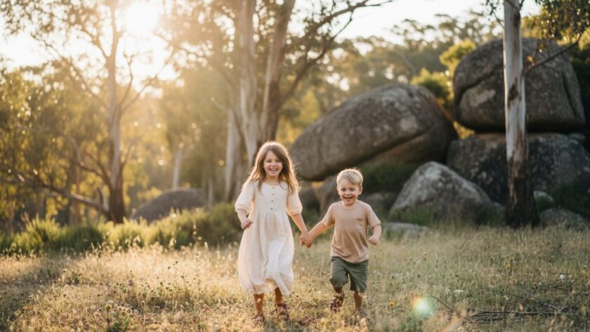 A wide-angle, sun-drenched photograph capturing an authentic kids photography Black Hill Victoria moment: two siblings laughing joyfully while running through a sunlit field with Black Hill's characteristic trees in the background, professional color grading enhancing the golden hour glow.