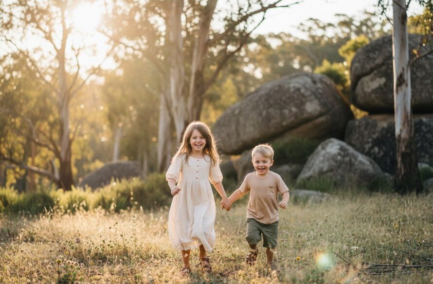 A wide-angle, sun-drenched photograph capturing an authentic kids photography Black Hill Victoria moment: two siblings laughing joyfully while running through a sunlit field with Black Hill's characteristic trees in the background, professional color grading enhancing the golden hour glow.