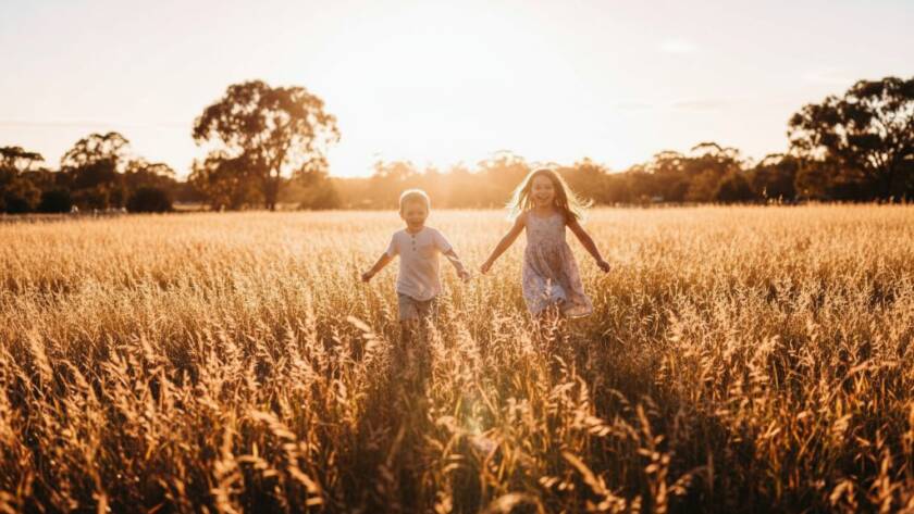 A wide shot capturing authentic Narre Warren kids photography: two joyful children, a boy and a girl, laughing as they run through a field of golden grass during sunset at Berwick Springs Reserve, Narre Warren. The dramatic backlighting creates a warm, cinematic glow, highlighting their playful silhouettes against the vibrant sky. Their faces are lit with pure happiness, embodying a precious, candid moment.