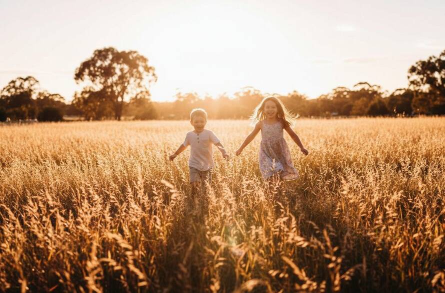 A wide shot capturing authentic Narre Warren kids photography: two joyful children, a boy and a girl, laughing as they run through a field of golden grass during sunset at Berwick Springs Reserve, Narre Warren. The dramatic backlighting creates a warm, cinematic glow, highlighting their playful silhouettes against the vibrant sky. Their faces are lit with pure happiness, embodying a precious, candid moment.