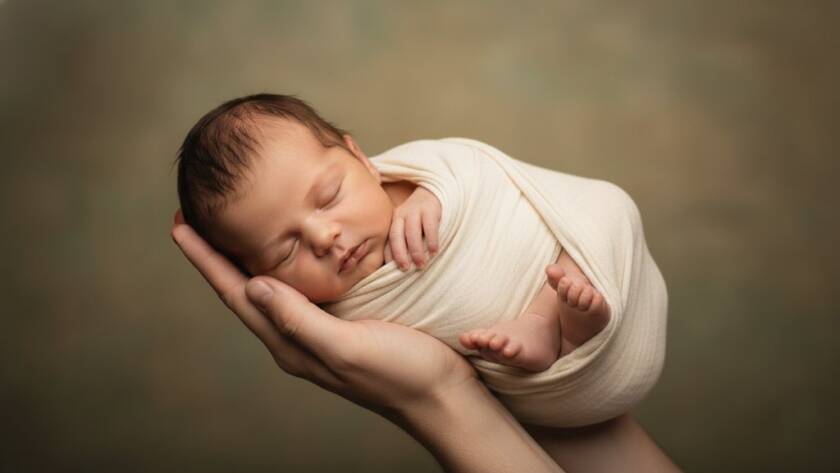 An intimate, beautifully lit studio portrait showcasing authentic newborn photography in Deer Park, Victoria, featuring a peacefully sleeping baby wrapped in soft fabric, held gently in a parent's hands, with warm, golden light creating a tender, ethereal glow.