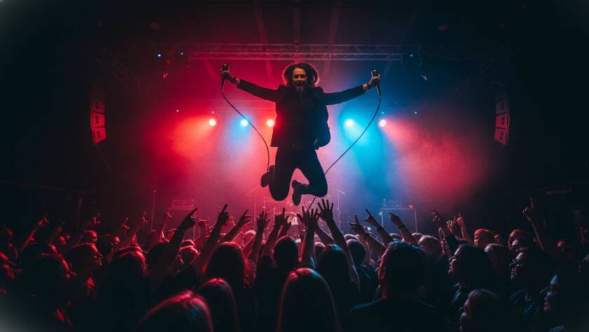 A wide-angle, low-angle professional photograph capturing a lead guitarist mid-shred under dramatic blue and red stage lights at a vibrant Beaconsfield live music event, with the crowd's hands raised in the foreground, showcasing powerful Beaconsfield live music photography dynamic shots.