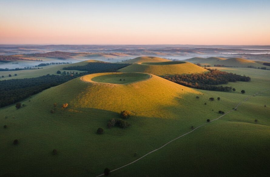 An awe-inspiring drone photograph capturing the expansive, verdant rolling hills and distant volcanic cones of Black Hill, Victoria at sunrise, with golden light illuminating the landscape and a misty valley below, showcasing the dramatic scale and beauty of the region from an epic high-angle panoramic view, highlighting the unique terrain for Black Hill Victoria drone photography panoramic views.