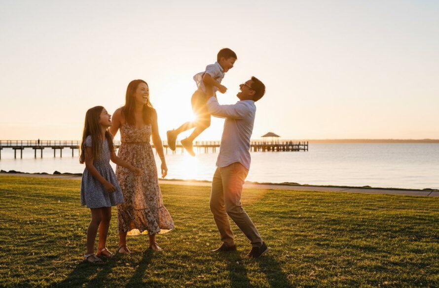 A joyful family laughing genuinely during candid Rippleside family photography Geelong, playing by the water's edge at sunset, with golden light reflecting off Corio Bay and the iconic Rippleside Park playground in the background. The father lifts a child high, capturing a perfect, unposed moment of pure happiness.