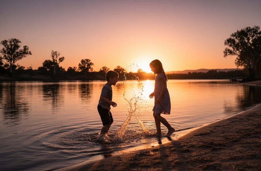 A professional, cinematic photograph capturing authentic kids photography moments Mildura riverfront, showing two children laughing joyfully while splashing in the golden light of sunset by the Murray River, their silhouettes vibrant against the water.