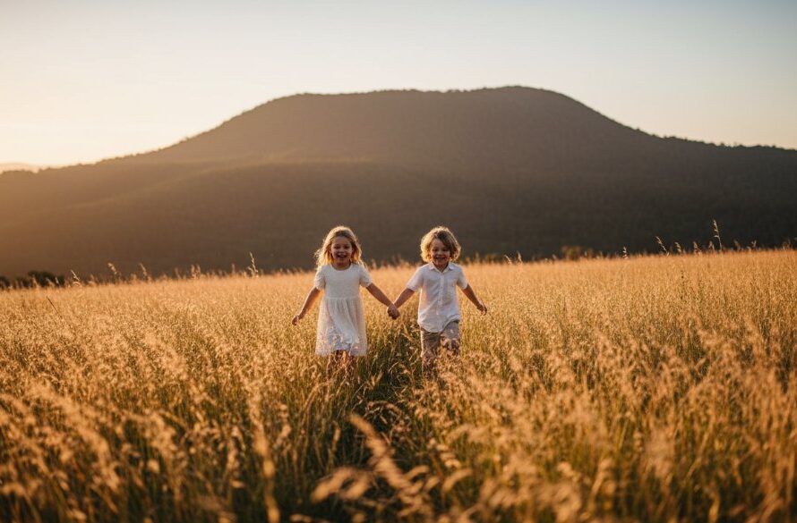 A candid, sun-kissed portrait of two laughing children running through a field of golden grass near Mount Macedon, captured as an authentic Macedon kids photography moment, with soft backlighting creating a dreamy, ethereal glow and a wide-angle lens for an epic sense of freedom and joy. The image is professionally color-graded with warm tones.