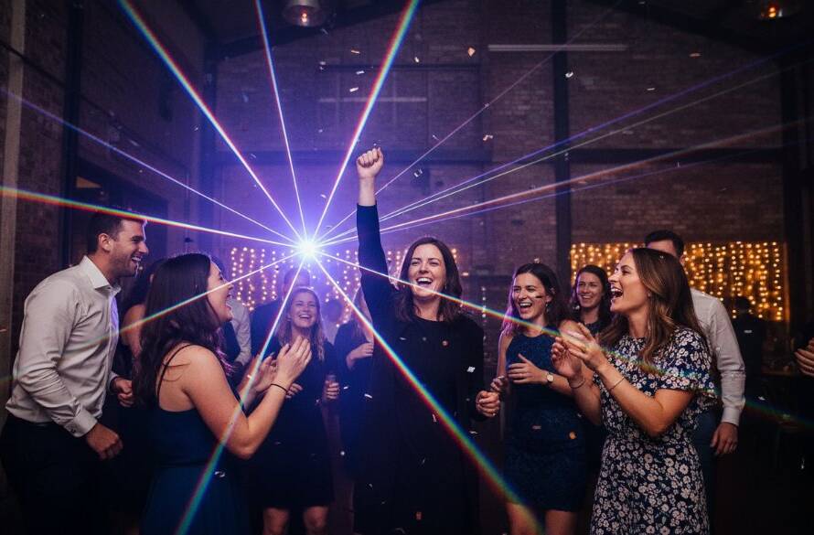 An emotionally charged, wide-angle shot Capturing Authentic Party Moments Brooklyn Victoria, featuring a group of friends cheering enthusiastically under dynamic, colourful stage lights at a lively community hall, capturing their genuine joy and connection with professional, cinematic colour grading.