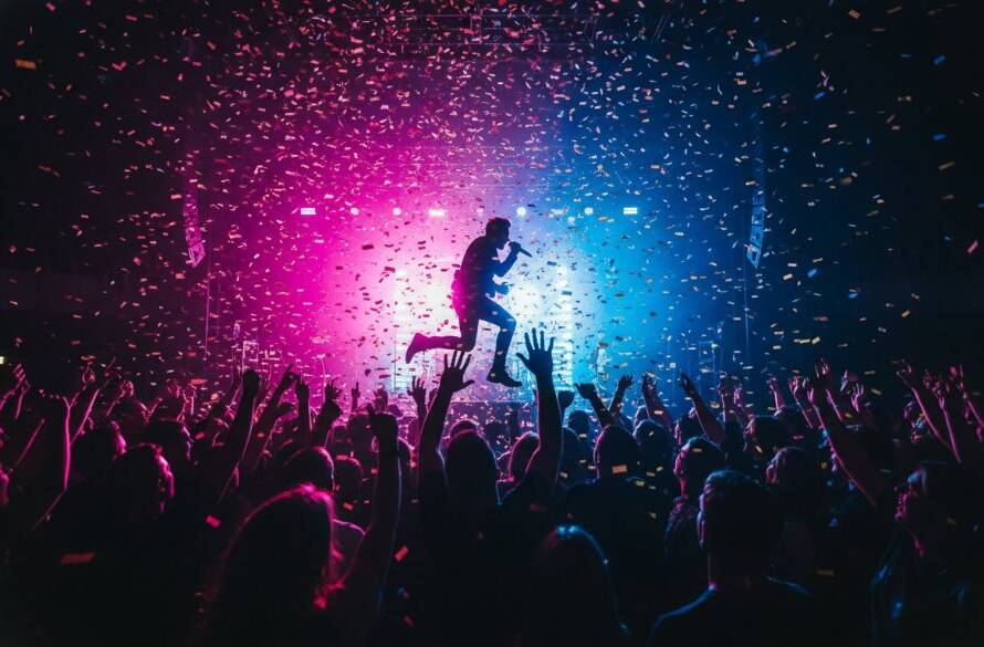 An electrifying shot showcasing the peak of a live performance, capturing Beaumaris live music event photography with a lead singer silhouetted by vibrant stage lights, confetti, and an ecstatic crowd at a Beaumaris venue.
