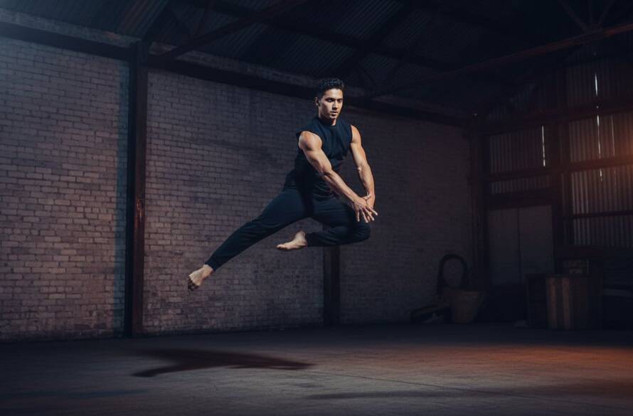 A male dancer in mid-air, performing an acrobatic leap during a dynamic dance photography moment in Clayton South, illuminated by dramatic stage lighting with an industrial urban backdrop.