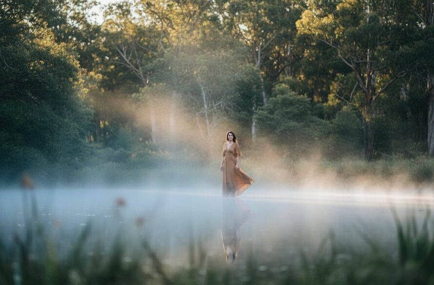 An epic, dramatic fine art photograph of a solitary woman in a flowing dress by the mist-shrouded mineral springs in Daylesford, Victoria, bathed in ethereal light, symbolising the emotional depth of capturing emotive fine art photographs in Daylesford's mineral springs.