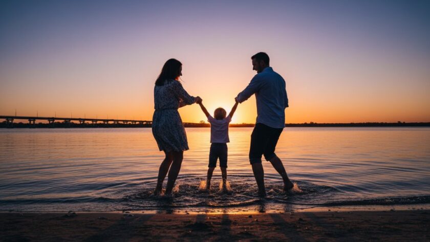A wide, cinematic shot capturing genuine Yarrawonga family moments: a family of three (parents and young child) laughing joyfully and splashing playfully at the golden hour on the sandy foreshore of Lake Mulwala, with the iconic Yarrawonga-Mulwala Bridge in the softly blurred background, silhouetted against a dramatic orange and purple sunset sky. Professional, color-graded, high-impact.