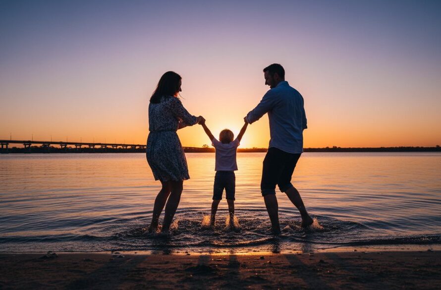 A wide, cinematic shot capturing genuine Yarrawonga family moments: a family of three (parents and young child) laughing joyfully and splashing playfully at the golden hour on the sandy foreshore of Lake Mulwala, with the iconic Yarrawonga-Mulwala Bridge in the softly blurred background, silhouetted against a dramatic orange and purple sunset sky. Professional, color-graded, high-impact.