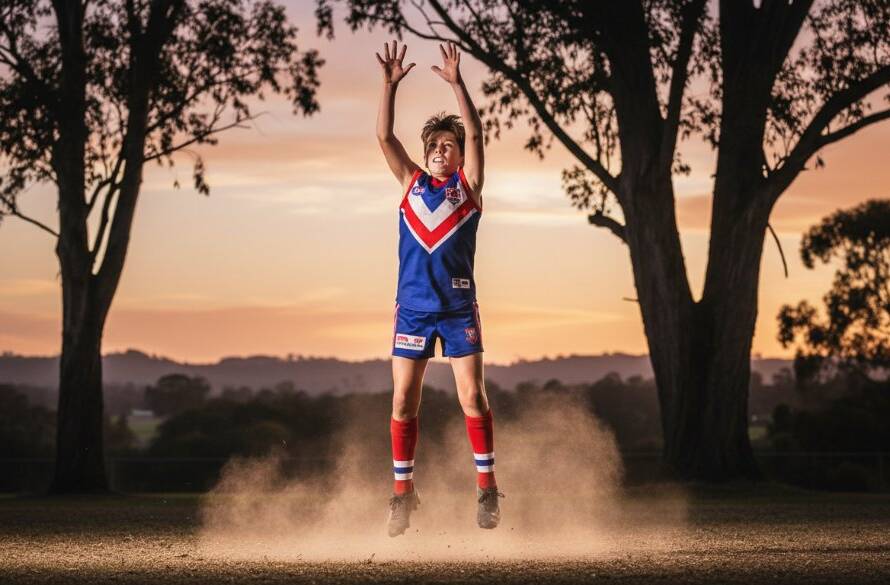Dynamic close-up shot of a young Aussie Rules footballer in mid-air, hands on the ball, during a crucial play, brilliantly capturing junior footy action Wonga Park. The player's determined expression and the blurred background convey intense focus and speed, illuminated by dramatic late afternoon sunlight, with green oval details.