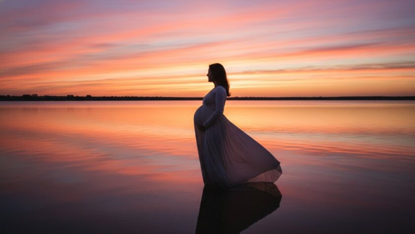 An epic, wide-angle professional photograph capturing a radiant pregnant woman silhouetted against a golden sunset over Lake Mulwala in Yarrawonga, Victoria, her hand gently cradling her belly, dramatic lighting creating a serene and timeless maternity portrait.