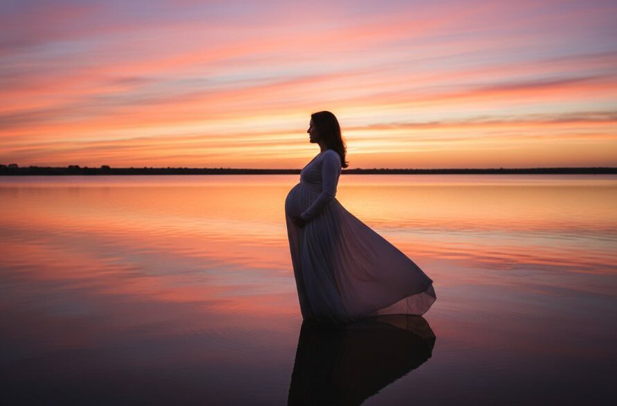 An epic, wide-angle professional photograph capturing a radiant pregnant woman silhouetted against a golden sunset over Lake Mulwala in Yarrawonga, Victoria, her hand gently cradling her belly, dramatic lighting creating a serene and timeless maternity portrait.