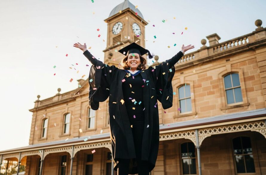 A jubilant graduate in their cap and gown, framed against the historic brickwork of the Castlemaine Gaol, tossing their mortarboard high into the golden hour sky, exemplifying Castlemaine graduation photography capturing authentic milestone moments, with dramatic lens flare and a shallow depth of field.