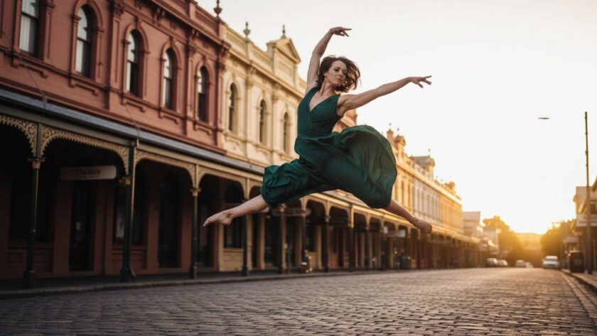 A ballet dancer in a stunning red costume performing an elegant leap mid-air, silhouetted against the iconic, golden-hued historic architecture of Clunes, Victoria, Australia, showcasing Clunes Heritage Dance Photography Victoria with dramatic light and shadow.
