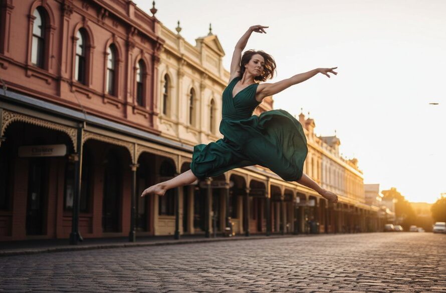 A ballet dancer in a stunning red costume performing an elegant leap mid-air, silhouetted against the iconic, golden-hued historic architecture of Clunes, Victoria, Australia, showcasing Clunes Heritage Dance Photography Victoria with dramatic light and shadow.