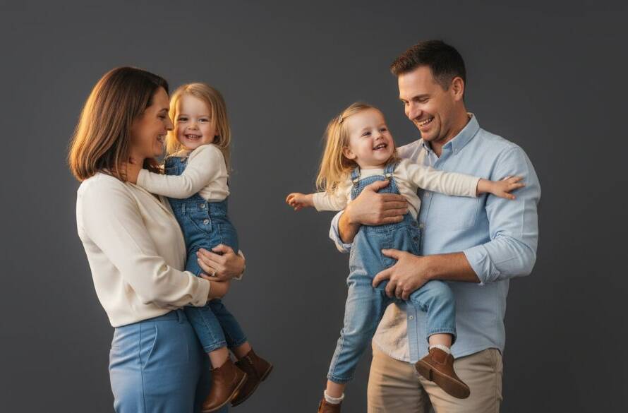 A breathtaking studio family portrait, an 'epic moment' showing a Narre Warren family of four laughing joyously, caught in dramatic, soft golden light against a deep grey backdrop. The parents are embracing their two children, one toddler looking up playfully, the other child with a radiant smile. This 'Crafting Timeless Studio Family Portraits Narre Warren' image highlights their authentic connection and happiness, with professional colour grading creating a warm, inviting atmosphere.
