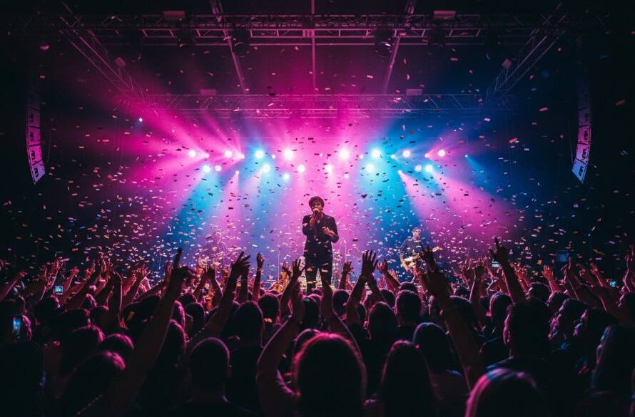 Epic wide shot of a band performing on stage at a vibrant Deepdene music venue, captured by Deepdene Live Gig Photography Melbourne. Dramatic stage lights silhouette the lead singer mid-scream, with a sea of enthusiastic audience members hands raised in the foreground, professional colour grading.