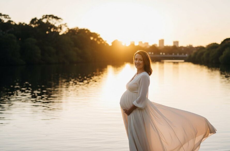 An expectant mother in a flowing gown, silhouetted against a dramatic sunset over the Barwon River in Drumcondra, Victoria, showcasing breathtaking Drumcondra Victoria waterfront maternity photography with professional lighting and warm tones.