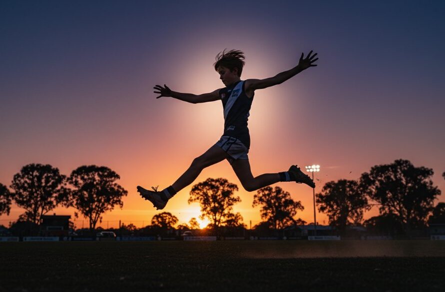 A thrilling moment of dynamic Bacchus Marsh junior sports photography, showing a young Australian Rules Football player in mid-air, kicking for goal under dramatic stadium lights at dusk.