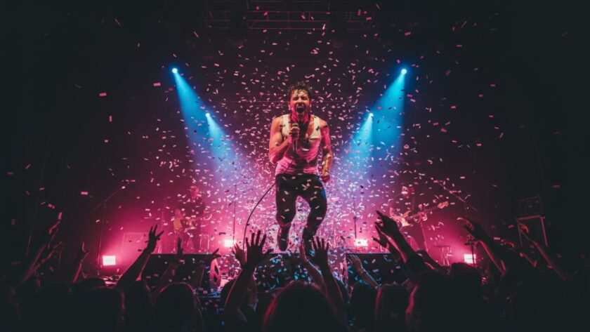 A vibrant, wide-angle photograph capturing the dynamic Traralgon concert photography services in action, showing a lead singer mid-scream under dramatic red and blue stage lights, with blurred audience hands in the foreground, conveying immense energy and an epic musical moment.