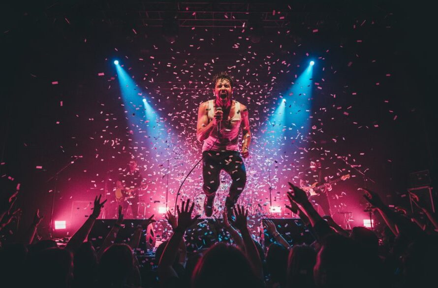 A vibrant, wide-angle photograph capturing the dynamic Traralgon concert photography services in action, showing a lead singer mid-scream under dramatic red and blue stage lights, with blurred audience hands in the foreground, conveying immense energy and an epic musical moment.