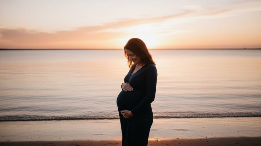 A radiant pregnant woman stands on the sandy shores of Corio Bay in East Geelong at sunset, her silhouette outlined by the golden light, showcasing an East Geelong maternity photoshoot bayside glow, as she gazes softly at her baby bump, with the calm water reflecting the dramatic sky.