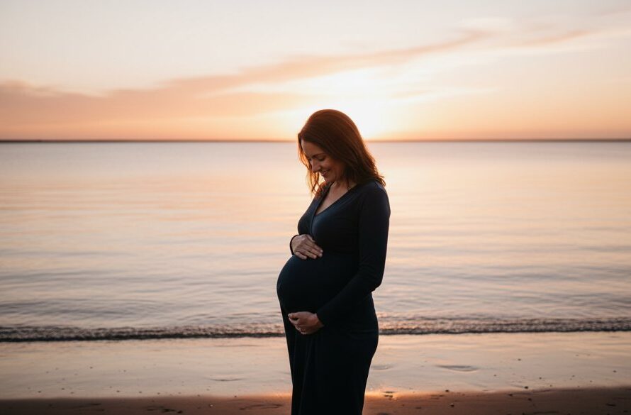 A radiant pregnant woman stands on the sandy shores of Corio Bay in East Geelong at sunset, her silhouette outlined by the golden light, showcasing an East Geelong maternity photoshoot bayside glow, as she gazes softly at her baby bump, with the calm water reflecting the dramatic sky.