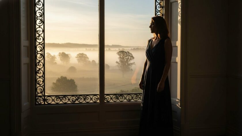 A stunning, cinematically lit 'epic moment' photograph of a woman gracefully posing by a large window overlooking a misty morning landscape in Wangaratta, capturing an empowering boudoir photography experience with dramatic shadows and golden hour glow.