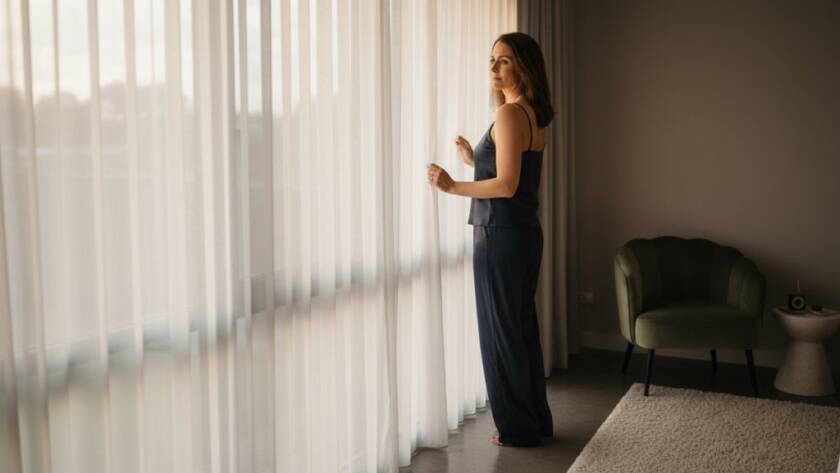 An empowering boudoir shoot Truganina: A confident woman gazing into the distance, framed by soft, dramatic light filtering through sheer curtains in a luxurious Truganina studio, capturing a moment of profound self-love and elegance.