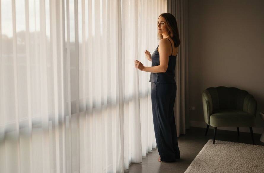 An empowering boudoir shoot Truganina: A confident woman gazing into the distance, framed by soft, dramatic light filtering through sheer curtains in a luxurious Truganina studio, capturing a moment of profound self-love and elegance.