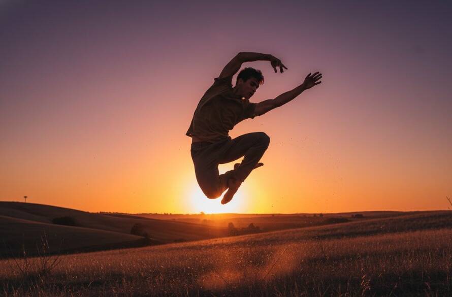 A male dancer in mid-air, striking a powerful pose against a dramatic sunset over rolling hills in Exford, Victoria, for an Exford dance photography outdoor portraiture session. Professional photography, dramatic lighting, rich colours.