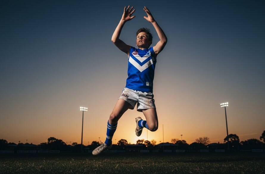 An expert sports photography Melton South junior footy moment: a young player in mid-air, kicking a football with intense focus, under dramatic stadium lights at sunset, capturing the raw emotion and athleticism.
