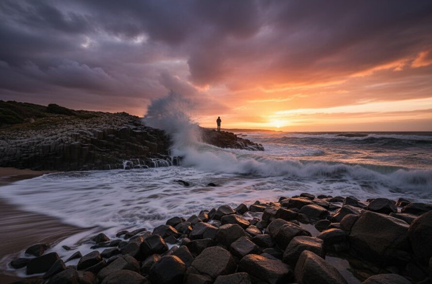 Dramatic long-exposure fine art photography Portland Victoria rugged coastline, featuring waves crashing against basalt columns at sunset, with a lone, silhouetted figure standing on a distant rock, evoking a sense of awe and connection to nature, professionally colour-graded.