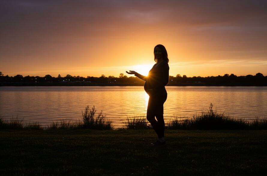 A radiant expectant mother, beautifully captured during a Footscray riverside maternity photoshoot experience, standing gracefully by the Maribyrnong River at sunset, dramatic backlighting illuminating her silhouette, showcasing the serene beauty of pregnancy.