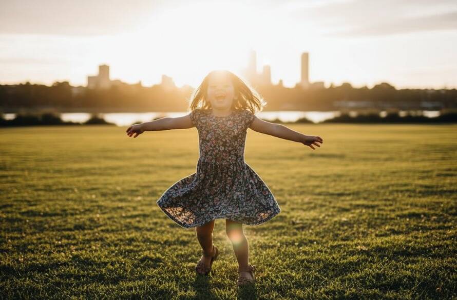A joyful child, around 5 years old, running barefoot through an open grassy field in Footscray, Victoria, during golden hour. The child is holding a brightly coloured kite that soars against the clear blue sky. Dramatic backlight creates a beautiful halo effect around their hair, casting long shadows. The setting includes distant, blurred Footscray cityscape elements and the Maribyrnong River. Professional, cinematic photography with warm, vibrant colour grading and shallow depth of field, capturing an epic moment of freedom and happiness. This image represents Footscray vibrant kids photography sessions.