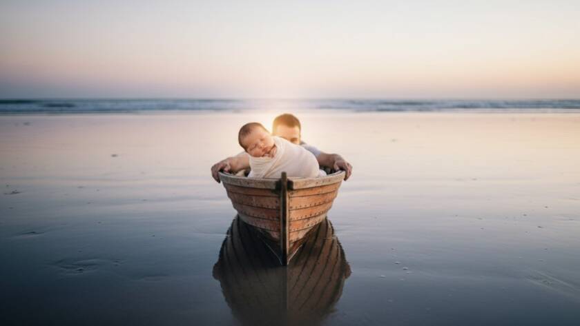 A heartwarming, softly lit, wide-angle photograph capturing a sleeping newborn baby nestled gently in a rustic wooden boat on a Seaford beach at dawn, with parents' hands protectively hovering, embodying heartfelt newborn photography Seaford Victoria coastal memories.