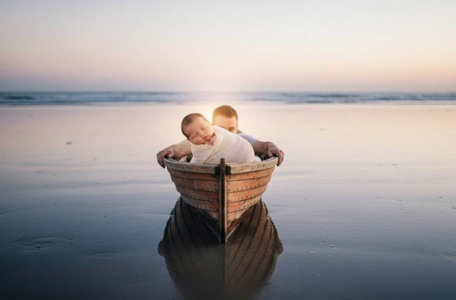 A heartwarming, softly lit, wide-angle photograph capturing a sleeping newborn baby nestled gently in a rustic wooden boat on a Seaford beach at dawn, with parents' hands protectively hovering, embodying heartfelt newborn photography Seaford Victoria coastal memories.